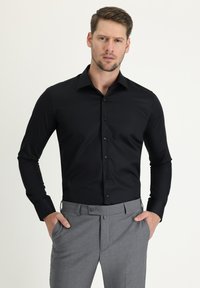 Camisa negra de manga larga con cierre de botones, con un diseño ajustado y textura suave, combinada con pantalones grises de corte a medida.