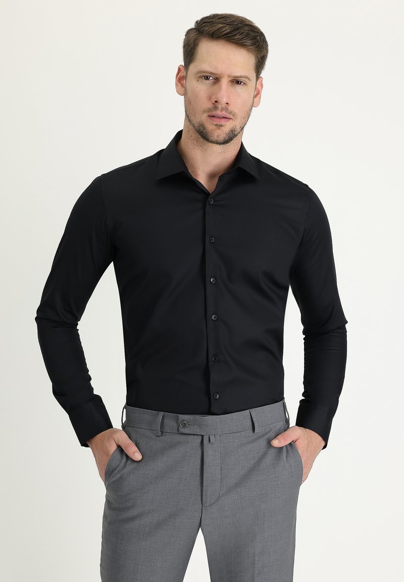 Camisa negra de manga larga con cierre de botones, con un diseño ajustado y textura suave, combinada con pantalones grises de corte a medida.