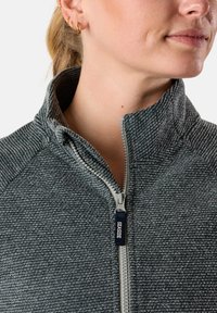 Grå zip-up jacka med texturerat stickat material, hög krage och en dragkedja med en blå etikett. Förstärkt med en diagonal söm.