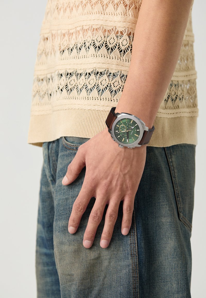 Montre chronographe avec un cadran vert, une lunette en argent et un bracelet en cuir brun ; portée sur une main contre un fond de haut en tricot beige.