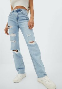 Mujer vistiendo jeans rectos desgastados de color azul claro, una camiseta corta blanca y zapatillas blancas, de pie frente a un fondo liso.