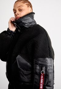 Zwarte schapenvachtjas met een hoge kraag, leren accenten, geribbelde manchetten en een ritszak met een rode label met de tekst "VERWIJDER VOORDAT."