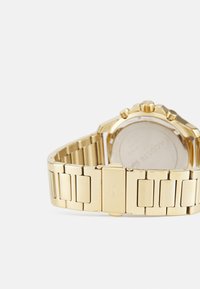 Reloj dorado con una correa metálica que presenta eslabones de diferente ancho, una caja redonda y tres pulsadores en el lateral, reflejando la luz.