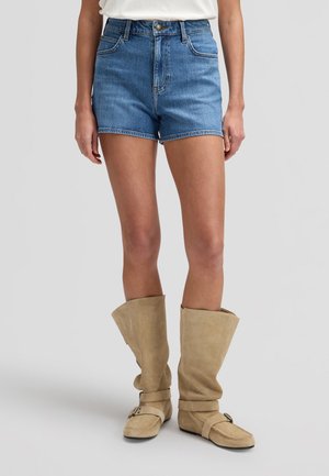 Person trägt blaue Jeans-Shorts mit mittlerer Taille und beige Wildlederstiefel bis zum Knie mit Schnallenriemen, steht vor einem neutralen grauen Hintergrund.