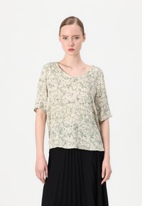 Lys grønn bluse med blomsterdesign, løs passform, korte ermene og V-hals. Har en teksturert stoff med svarte detaljer. Brukt sammen med en svart plisserte skjørt.