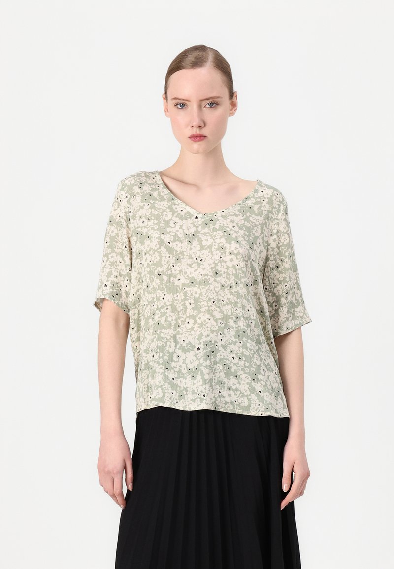 Lys grønn bluse med blomsterdesign, løs passform, korte ermene og V-hals. Har en teksturert stoff med svarte detaljer. Brukt sammen med en svart plisserte skjørt.