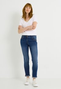 Jeans ajustados azul claro con un sutil desgaste, que cuentan con cinco bolsillos y costuras marrones, combinados con una camiseta blanca de manga corta y cuello en V, y zapatillas blancas.