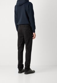 Černé kalhoty vyrobené z odolného materiálu, rovného střihu, s dvěma zadními kapsami na knoflíky, nošeny s tmavě modrou mikinou a tmavými botami.