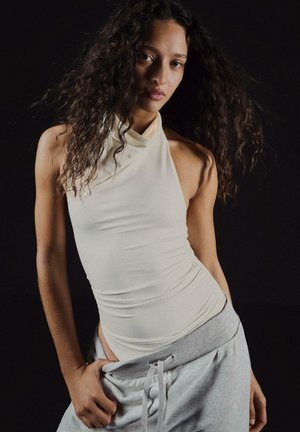 Junge Frau mit langen lockigen Haaren, die ein ärmelloses beiges Oberteil und graue Hose trägt, posiert vor einem schwarzen Hintergrund.