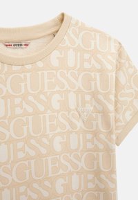 T-shirt beige con un motivo a logo "GUESS" ripetuto. Presenta un colletto a costine e un piccolo badge a triangolo con il logo sul petto.