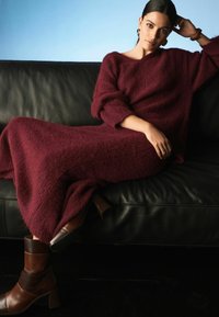 Burgundské pletené šaty s dlouhými rukávy a volným střihem. Hnědé kožené kotníkové boty na blokovém podpatku. Pozadí tvoří černá kožená pohovka.