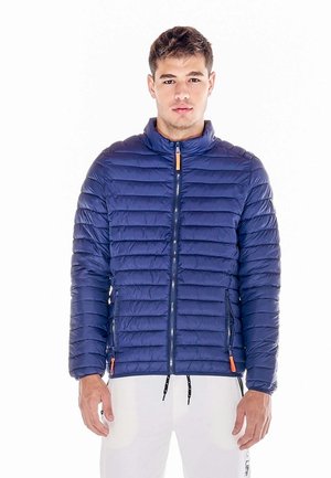 Un homme se tient sur un fond blanc, vêtu d’une veste matelassée bleu marine avec des tirettes de fermeture éclair orange et d’un pantalon blanc.