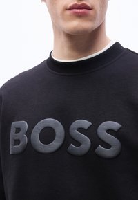 Κοντινό πλάνο ενός ατόμου που φοράει μαύρο φούτερ με μεγάλ α, ανάγλυφα γράμματα που σχηματίζουν τη λέξη "BOSS" στο στήθος.