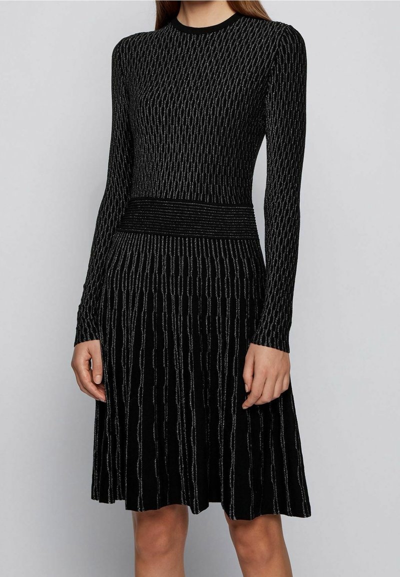 Femme portant une robe en maille noire à manches longues avec des rayures verticales argentées et une taille ajustée, debout devant un fond uni.
