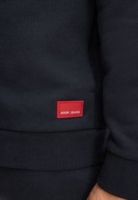 Felpa blu navy con texture a costine, dotata di un piccolo etichetta rossa con "JOOP! JEANS" vicino all'orlo. Design semplice, senza motivi visibili.