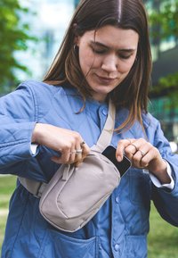 Borsa a spalla in tessuto beige con una tasca frontale. Persona che inserisce uno smartphone. Giacca blu trapuntata con bottoni tondi e tasche.