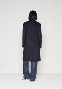 Marineblå lang uldfrakke med en lige silhuet, enkle knapdetaljer og knaplukning; parret med brede denimbukser og sneakers.