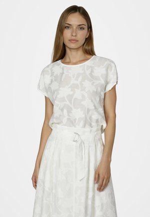 Frau trägt eine weiße Kurzarmbluse mit Blumenmuster und einen passenden hoch taillierten Rock mit Stoffgürtel und silberner Schnalle.