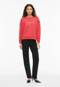 Sudadera roja con texto "imagine" en blanco, combinada con pantalones negros de pierna recta y tacones negros de punta puntiaguda. Tela suave, ajuste relajado.