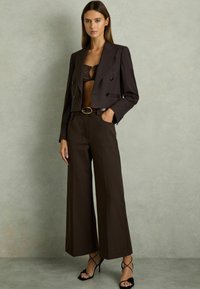 Bruin, dubbelgebreid cropped blazer, bijpassende high-waisted wijde broek, een bruine riem met gouden gesp en zwarte, gekruiste hoge hakken.