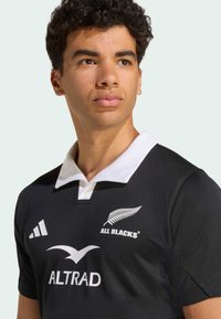 Schwarzes Rugbyshirt mit weißem Kragen, darauf das Logo "ALL BLACKS" und der Sponsor "ALTRAD" in Weiß. Kurze Ärmel, strukturierter Stoff.