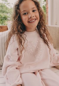 Felpa rosa chiaro con un'applicazione a forma di cuore in paillettes, abbinata a pantaloni coordinati. Tessuto morbido e strutturato con maniche a sbuffo.