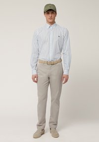 Lichtblauw gestreept overhemd met knopen, beige chino's, tan riem en olijfgroene pet; zachte textuur. Schoenen zijn lichtgrijze espadrilles.