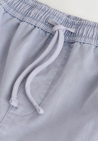 Pantalon à taille élastique bleu clair avec un cordon tressé assorti noué en un simple nœud sur un fond uni.