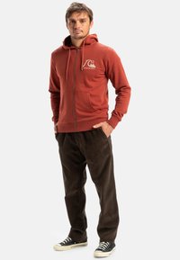 Red zip-up hoodie with a front pocket, featuring a logo on the chest, paired with brown corduroy pants and black sneakers.