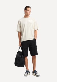 Camicia beige a maniche corte con tasca nera a contrasto, abbinata a pantaloni cargo neri. Porta una borsa a tracolla nera e indossa scarpe da ginnastica con motivo.