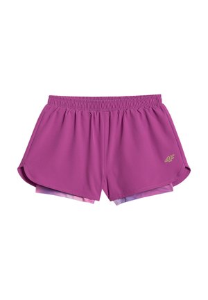 Młoda dziewczyna stoi, ubrana w różowo-fioletową koszulkę z motywem tie-dye, fioletowe szorty, białe skarpetki i czarne buty sportowe na jednolitym tle.
