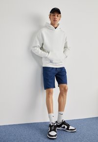 Jeune homme portant un sweat à capuche blanc, un short en jean bleu, une casquette noire, des chaussettes blanches Nike et des baskets Nike noir et blanc, debout contre un mur blanc.
