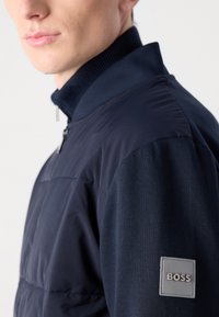 Navy blue jacket with a high collar, zip closure, and textured fabric. Features a gray patch with "BOSS" logo on the left sleeve.