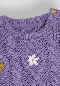 Lila stickad tröja med ett texturerat kabelmönster, en liten vit broderad blomma och träknappdetalj vid kragen.