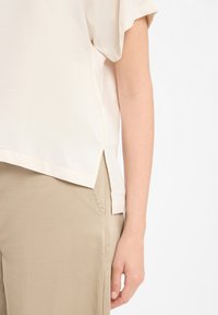 Licht beige T-shirt met korte mouwen en een ontspannen pasvorm, zijsplitten aan de onderzijde, gemaakt van gladde stof, gecombineerd met licht beige broeken.