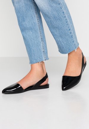 Voeten in lichtblauwe cropped jeans en zwarte gepatineerde slingback ballerina's met puntige neuzen op een gladde witte vloer.