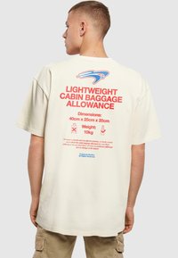 Beige oversized t-shirt med rød og blå tekst på ryggen, der beskriver tilladt håndbagage, dimensioner og vægtspecifikationer.
