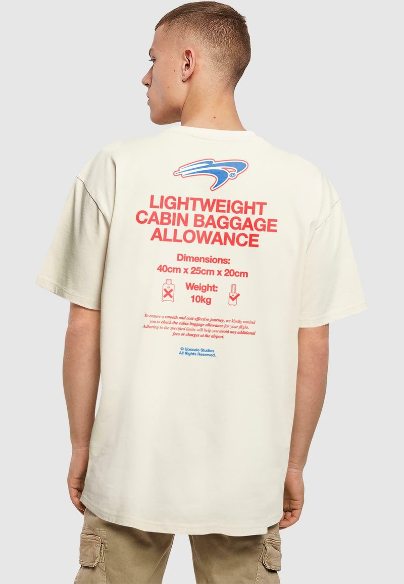 Beige oversized t-shirt med rød og blå tekst på ryggen, der beskriver tilladt håndbagage, dimensioner og vægtspecifikationer.