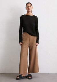 Zwarte lange mouw top, bruine wijde broek, en zwarte gestructureerde sandalen, tegen een neutrale achtergrond. Gestructureerde brei- en gladde stof.