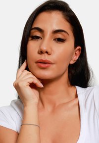 Primo piano di una persona con un top a coste bianco. Presenta lunghi capelli scuri, pelle liscia e un bracciale in argento al polso.