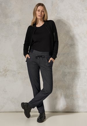 Veste zippée noire par-dessus un t-shirt noir, accompagnée d'un pantalon de jogging gris foncé avec poches et cordon de serrage, complété par des bottes noires.