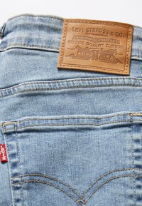 Tela de mezclilla azul claro con detalles de costura. Presenta un parche de cuero marrón con texto y gráficos grabados en la cinturilla. Etiqueta roja visible.