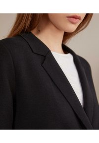 Černý přiléhavý sako s texturovanou látkou, s klopami do V a čistými liniemi, nošené přes bílý top. Detailní pohled.