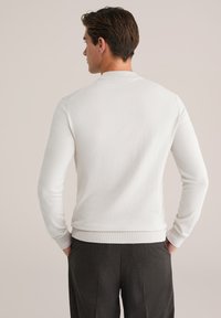 Valge, pika varrukaga sviiter ribatud krae ja mansettidega, millel on sile tekstuur ja keha lähedane disain. Kandmisel hallide pükstega.