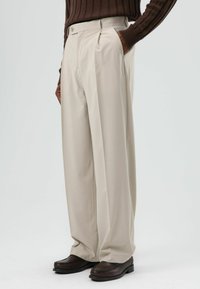 Beige, wijde broek van gladde stof met plooien aan de voorkant en zijzakken, gecombineerd met donkerbruine loafers.