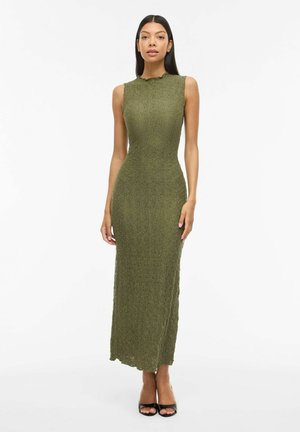Femme portant une robe midi texturée sans manches vert olive et des talons ouverts noirs, debout devant un fond blanc uni.