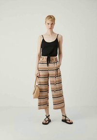 Schwarzes Tanktop mit dünnen Trägern, kombiniert mit gemusterten, weiten Hosen in Pfirsich, Schwarz und cremefarbenem Zickzack. Accessoires: kleine, tanfarbene Tasche.
