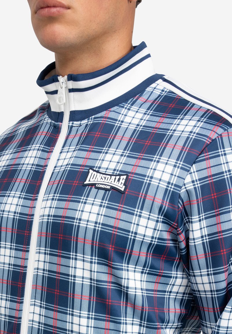 Karierte Zip-Jacke in Blau, Rot und Weiß. Sie verfügt über einen hohen Kragen, gestreifte Abschlüsse und ein gesticktes Logo auf der linken Brust. Glatte Textur.
