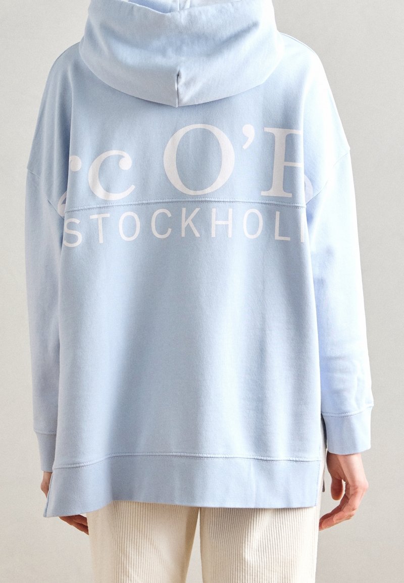 Personne portant un sweat à capuche bleu clair avec "STOCKHOLM" et un texte blanc partiel dans le dos, associé à un pantalon beige.
