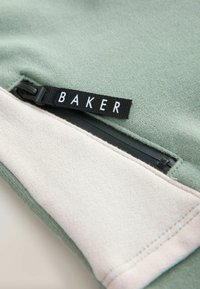 Zielono-beżowa tkanina z czarnym zamkiem błyskawicznym i czarną pętelką z napisem "BAKER" białymi literami.